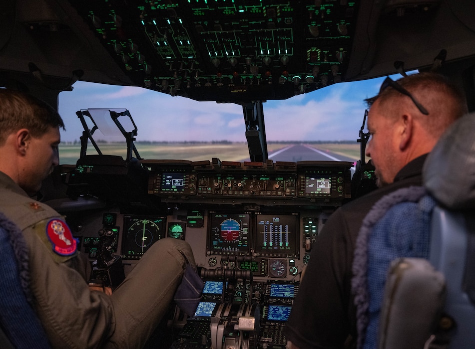 U.S. Air Force Maj. John Turbyfill, 58th Airlift Squadron assistant operations officer, left, leads Daniel Gray, Air Mobility Command (AMC) civic leader, right, through a C-17A Flight Simulator, known as a Weapon System Trainer, at Altus Air Force Base, Oklahoma, April 9, 2026. AMC senior leaders and civic leaders had the opportunity to operate a flight simulator which is used to train students at Altus. (U.S. Air Force photo by Airman 1st Class Emma Wright)