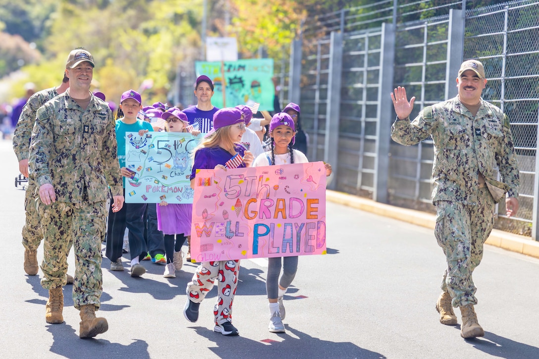 A large group of military children wearing purple clothing and hats walk in a parade with service members while carrying homemade signs.