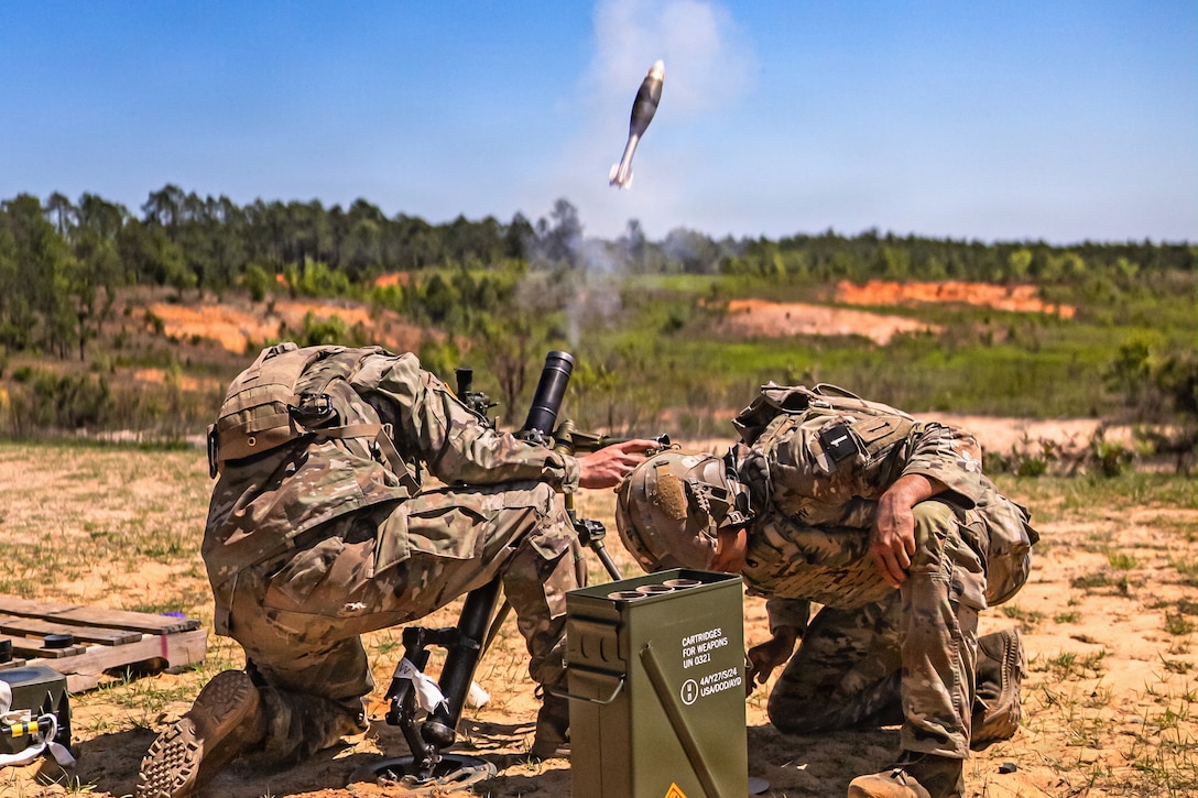 Two kneeling soldiers in tactical gear bend over in a field after firing a mortar into the air during the day.