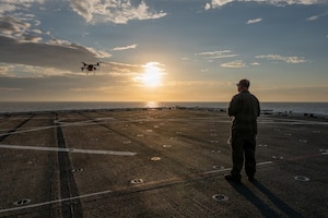 As the sun rises, a man flies a drone from a military ship in the water.