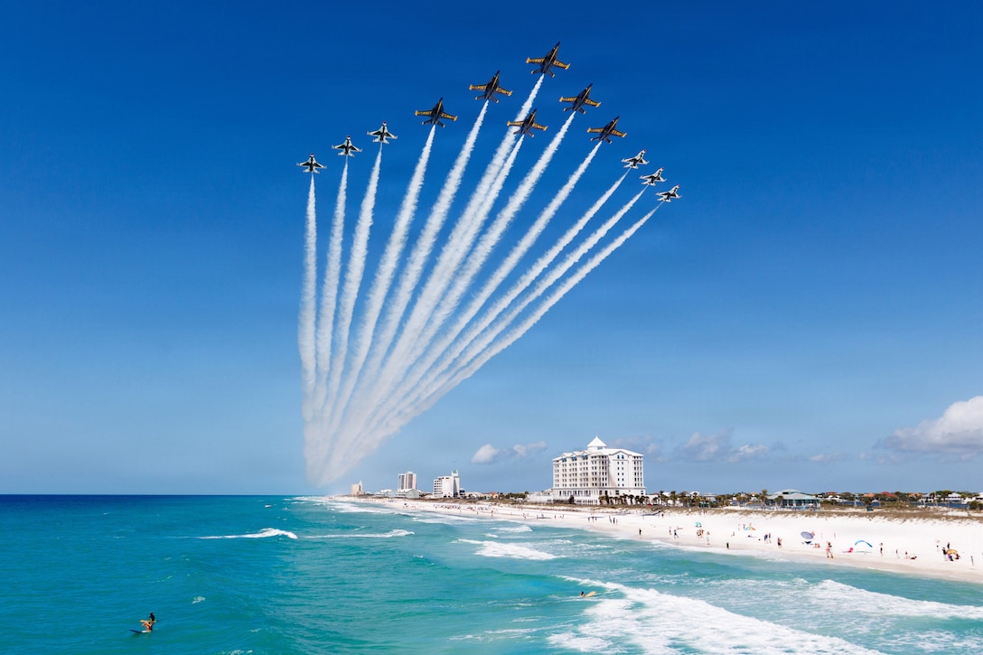 Twelve small airplanes fly in a V-shaped, triangular flight pattern above white sand and turquoise water, as people on the beach look up.