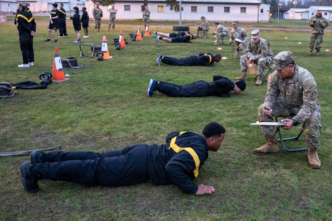 Five people lay facing the ground with their heads lifted slightly, arms bent and hands on the ground while other people in camouflage sit in front of them on chairs and hold clipboards.