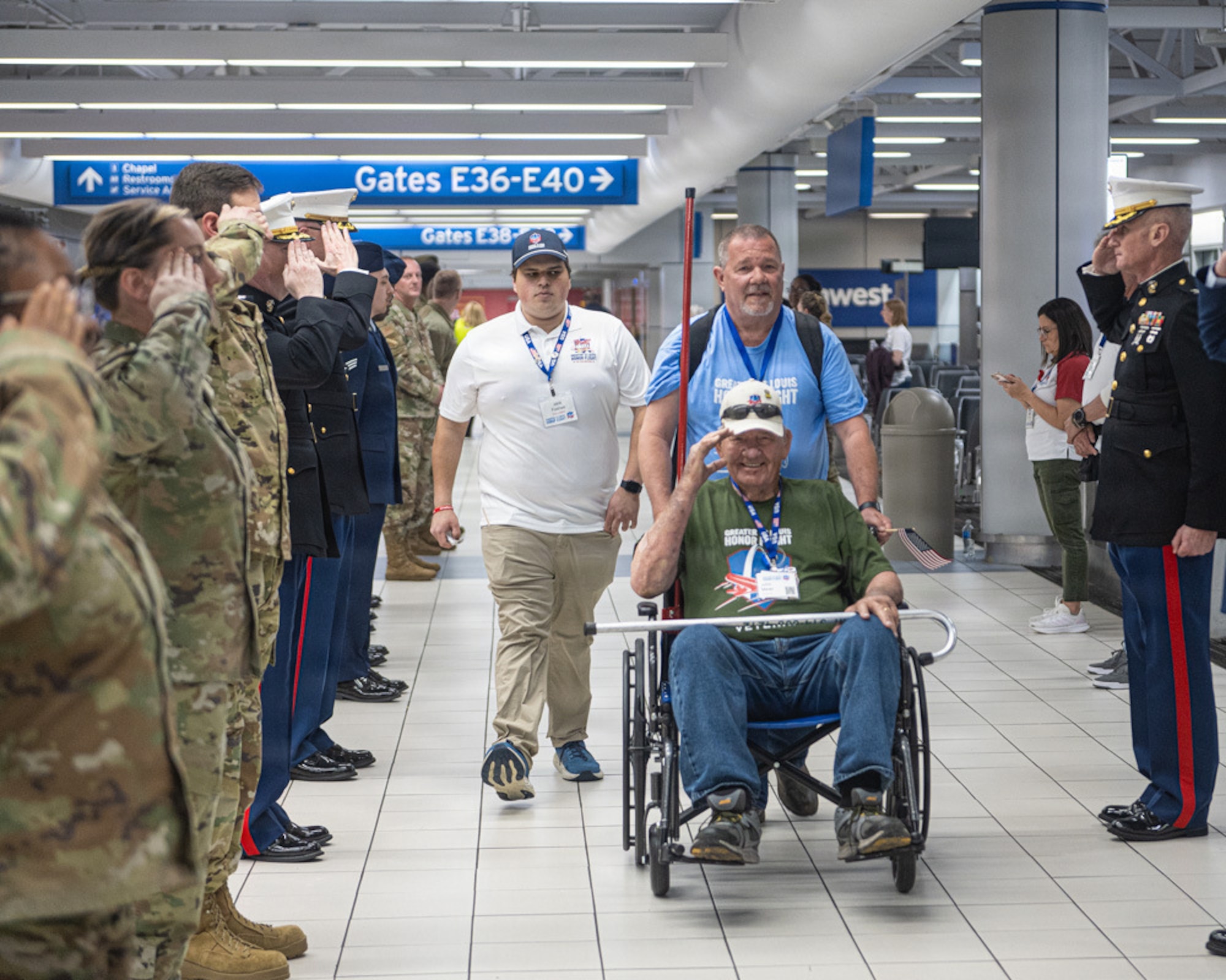 A veteran in a wheel chair salutes as he is wheeled past a line of servicemembers on each side of him.