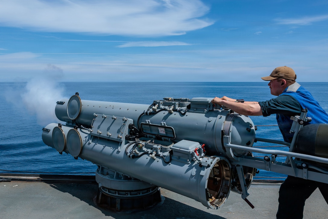 A person stands behind a large cannon-like structure attached to the edge of a ship's deck, with white smoke, blue water and a blue sky with clouds in the background.