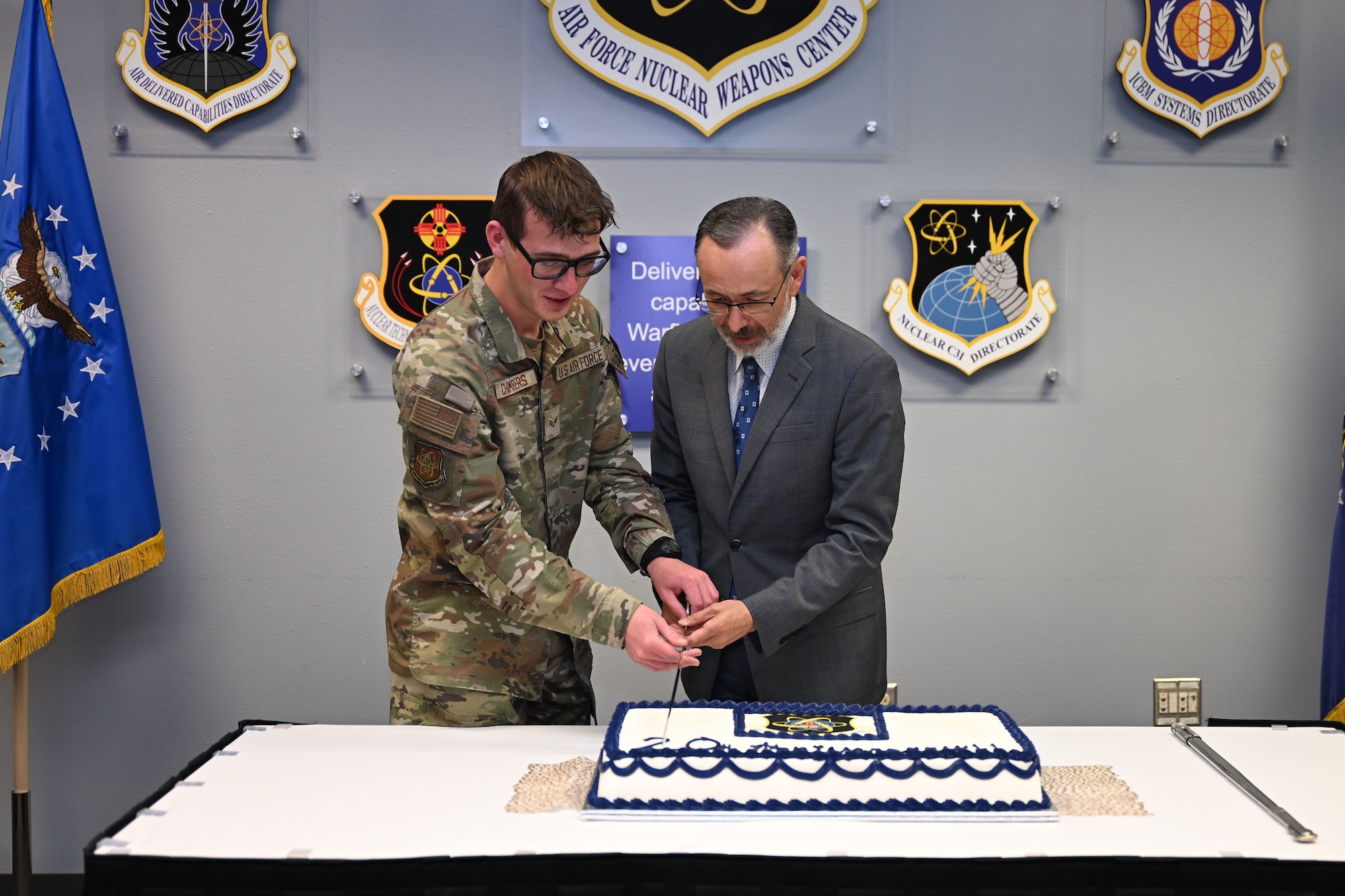 One man in uniform and one man in a suit use a sword to cut a cake