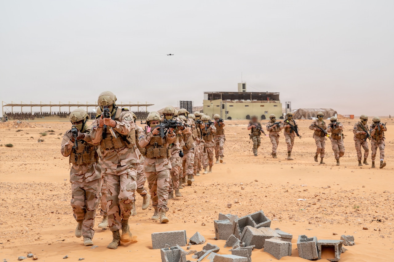 More than a dozen people in camouflage military uniforms aim weapons and walk through a desert.