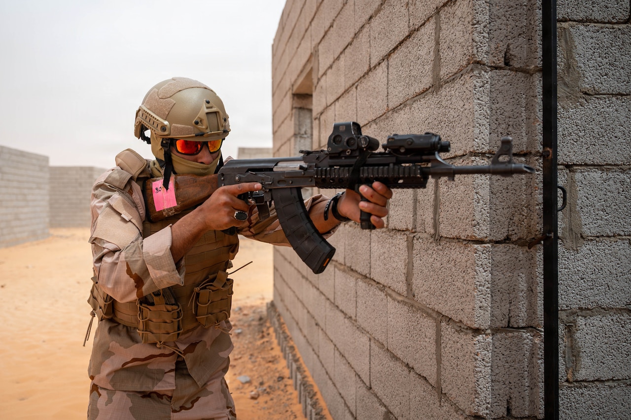 A person in a camouflage military uniform, helmet, goggles and face mask aims a weapon around the corner of a concrete building outside in a desert.