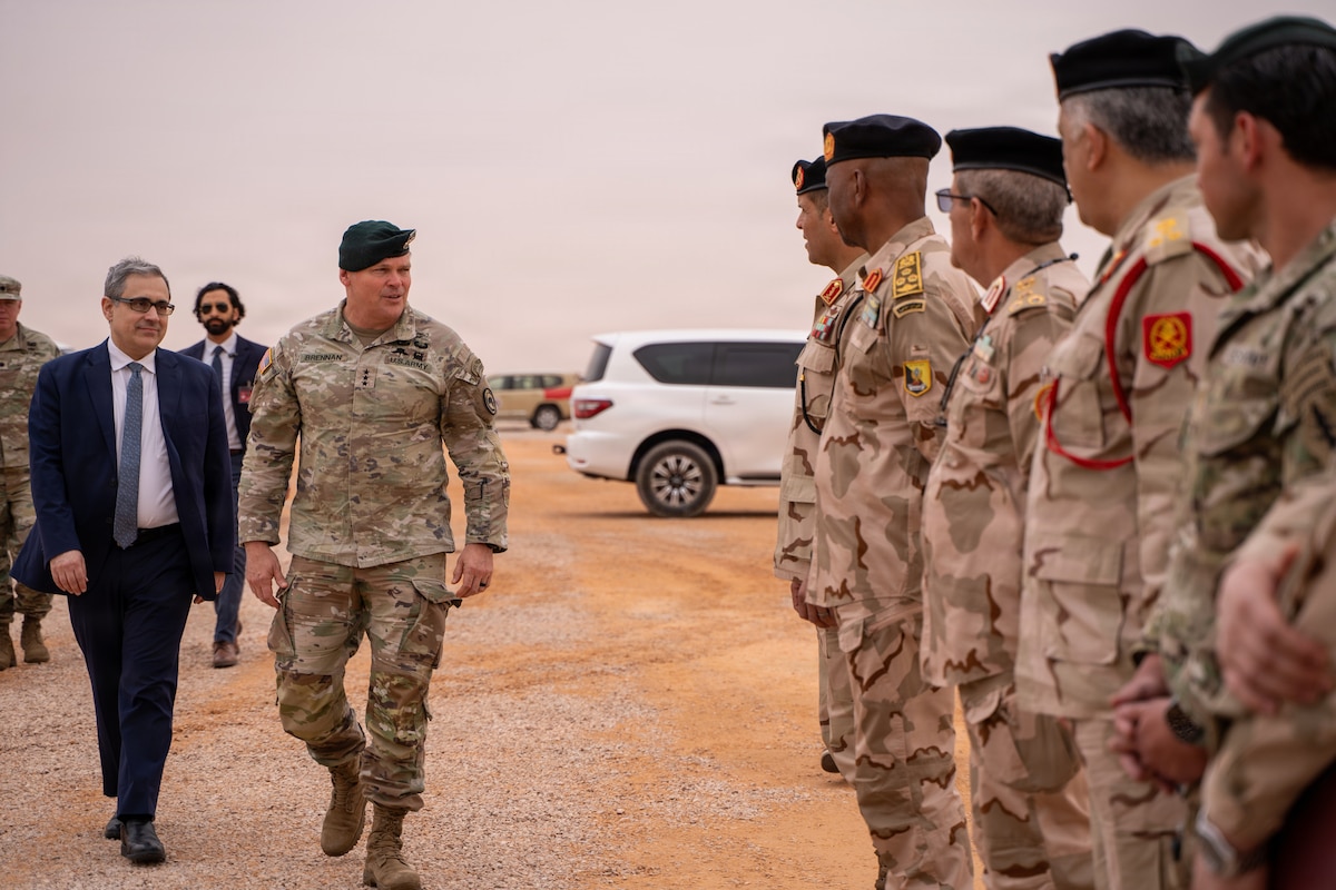 A man in a camouflage military uniform walks past a line of people in similar attire outside in a desert; to his right are two other men in business attire, and behind him are vehicles.