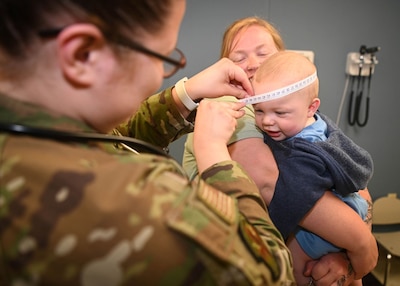 Medical professional in uniform uses a tape measure on a baby's head while a woman holds the baby.