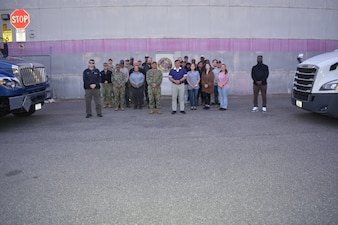 The NMRLC Transportation Directorate team members stand between two of the command's 18-wheeler vehicles that are central to its global logistics operations. The blue tractor is the International MV Series and the white one is the Freightliner Cascadia.