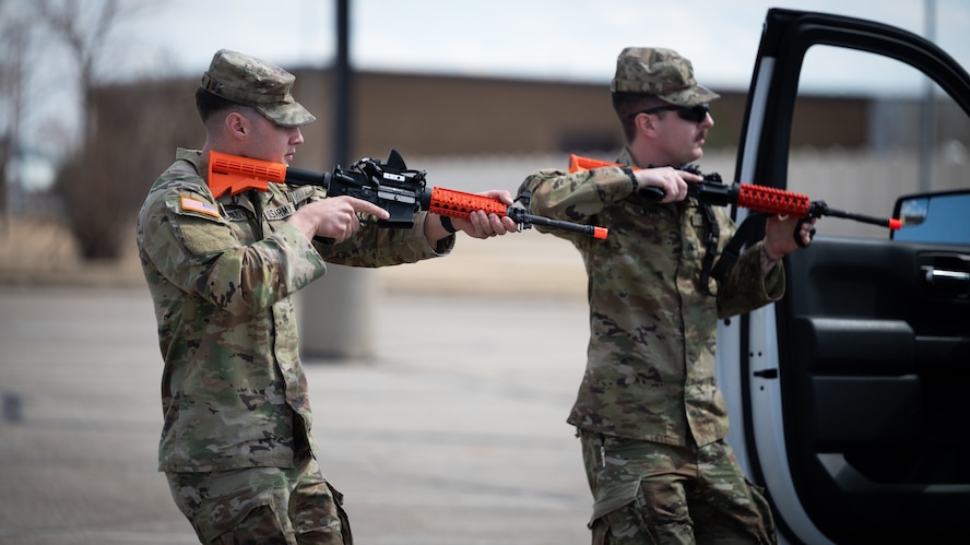 Two soldiers wielding training rifles approach a vehicle during a training exercise.