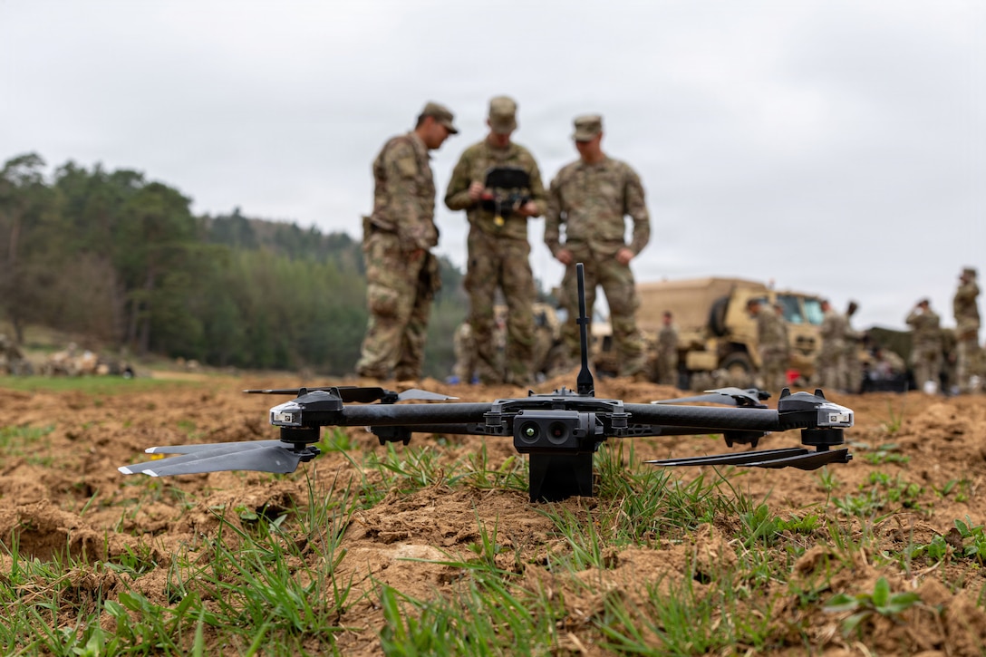 A close-up view of a drone sitting in a grassy, dirt terrain with three people in camouflage military uniforms standing in the background, holding the controls. In the distance, people in similar attire perform similar activities.