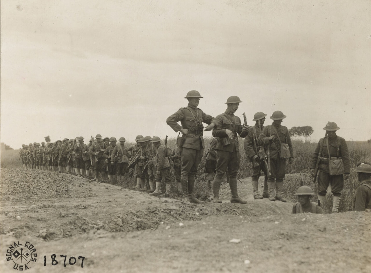 Dozens of men in military uniforms holding guns stand in formation in a dirt field as three other men in similar attire stand in a trench.