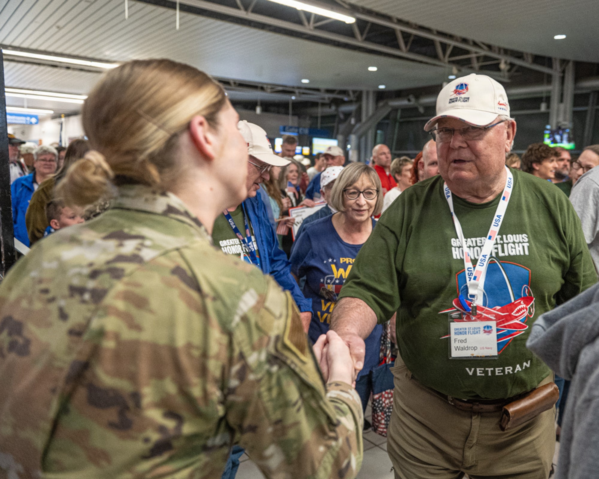 An Airmen to the left dress in camouflage shakes hands with a veteran with a green shirt and a white hat.