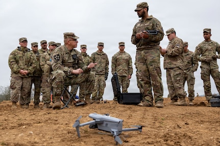 A man in a camouflage military uniform sits in a chair outside and speaks to a man in similar attire as a dozen other people, also in camouflage military uniforms, watch behind them. In the foreground, a drone sits in the dirt.