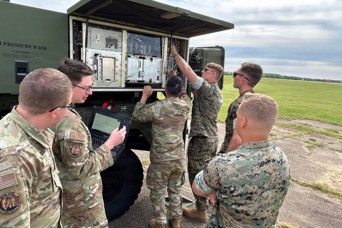 Two people in camouflage military uniforms work on a large air traffic control system outside while four other people in similar attire observe; one of them is holding up a computer for another person to look at.