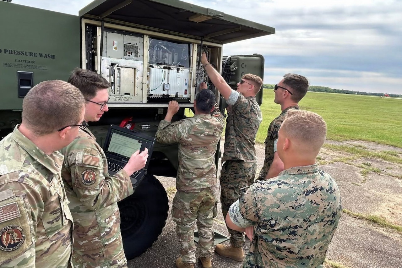 Two people in camouflage military uniforms work on a large air traffic control system outside while four other people in similar attire observe; one of them is holding up a computer for another person to look at.