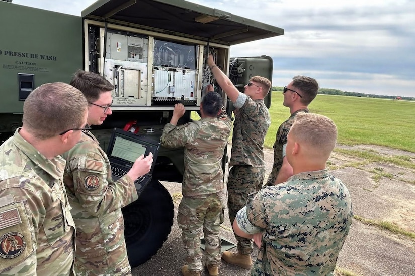 Two people in camouflage military uniforms work on a large air traffic control system outside while four other people in similar attire observe; one of them is holding up a computer for another person to look at.