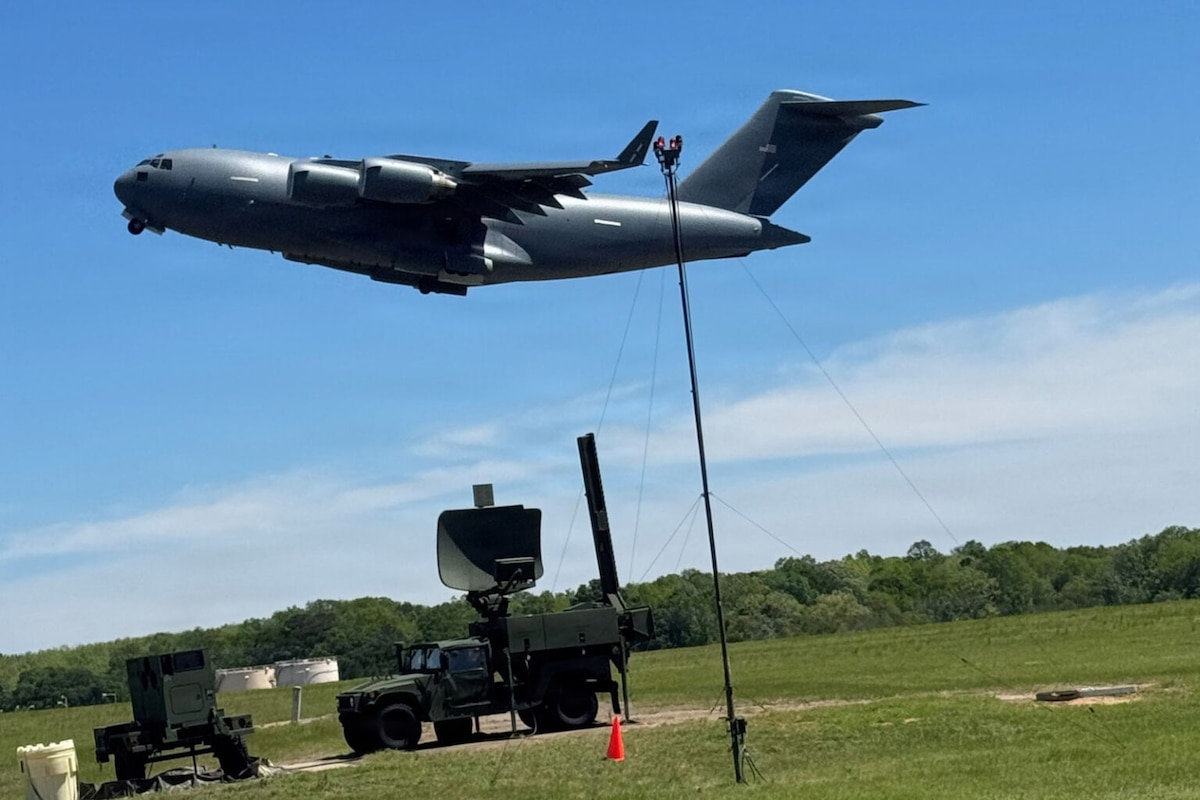 A large military aircraft flies over an air traffic control system attached to a military vehicle parked in a field.