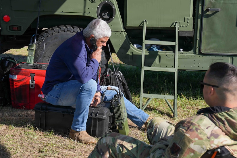 A man in casual attire talks on a radio while sitting on a black case outside next to a large air traffic control system; another man in a camouflage military uniform sits in a chair looking at the man talking on the radio.