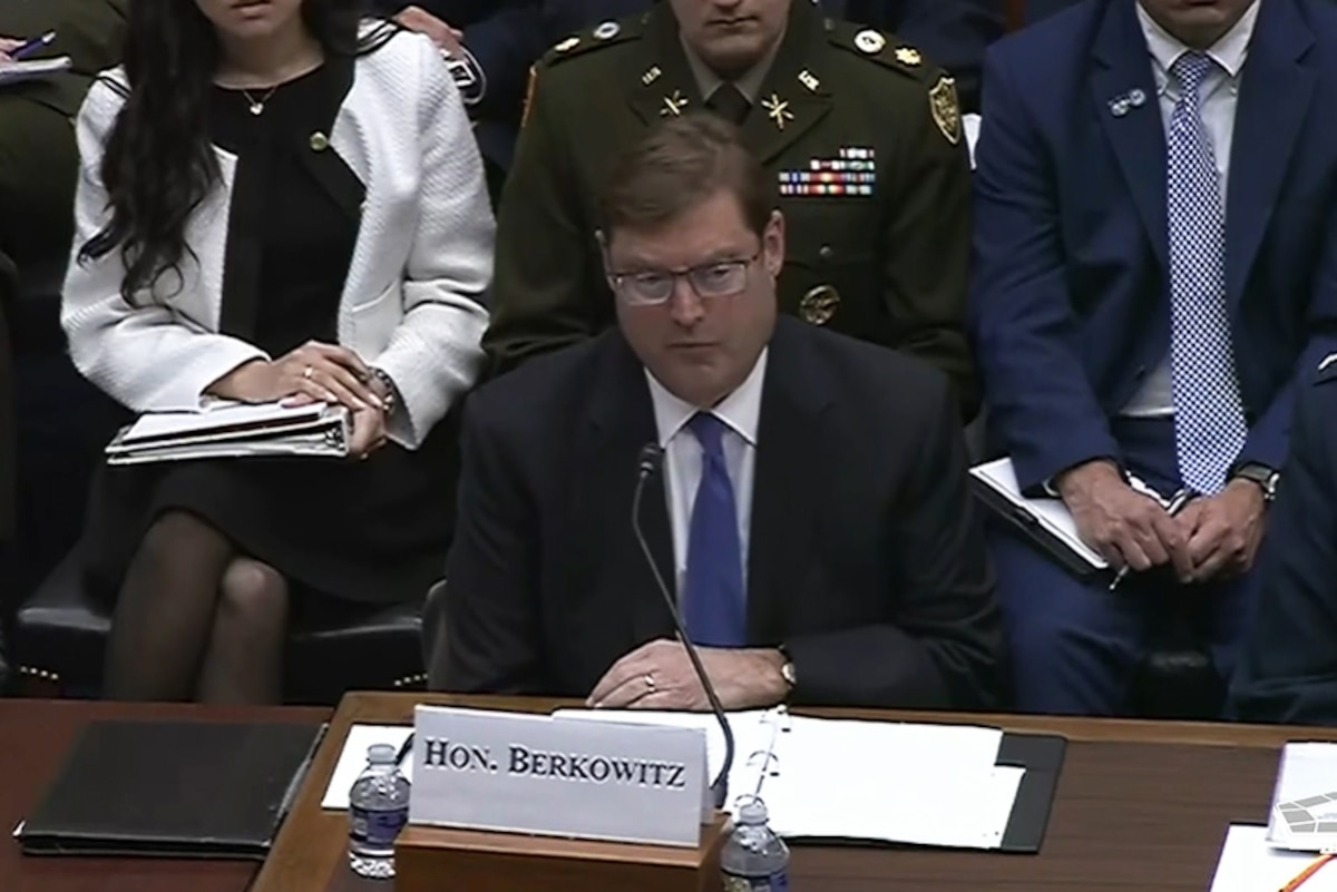 A man in business attire sits at a table and speaks into a microphone; people in similar attire are sitting behind him.