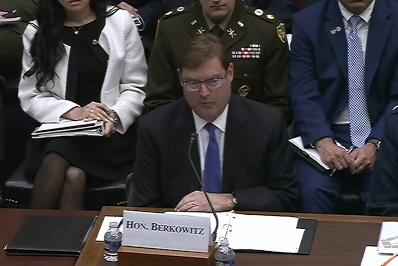 A man in business attire sits at a table and speaks into a microphone; people in similar attire are sitting behind him.