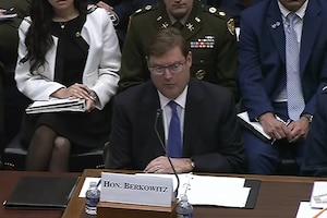 A man in business attire sits at a table and speaks into a microphone; people in similar attire are sitting behind him.