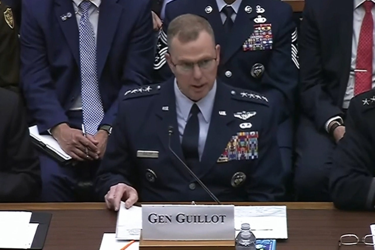 A man in a formal military uniform sits at a table and speaks into a microphone.
