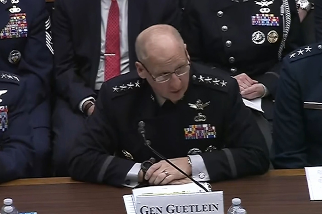 A man in a formal military uniform sits at a table and speaks into a microphone.