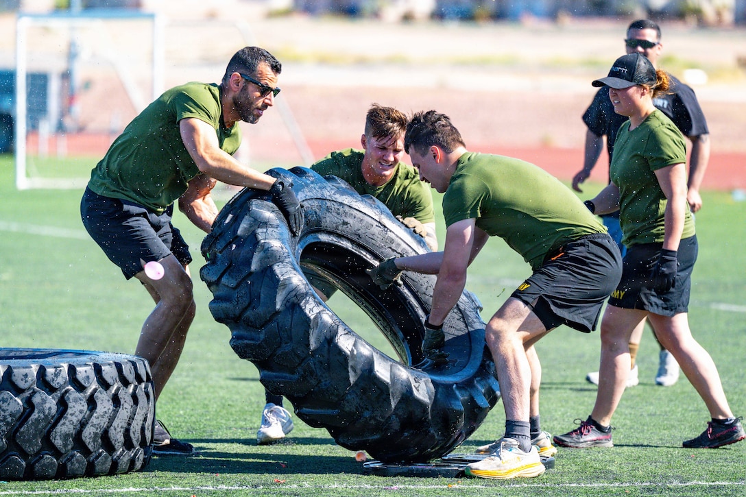 Four airmen in green shirts work together to move a large tire on a turf field during the day.