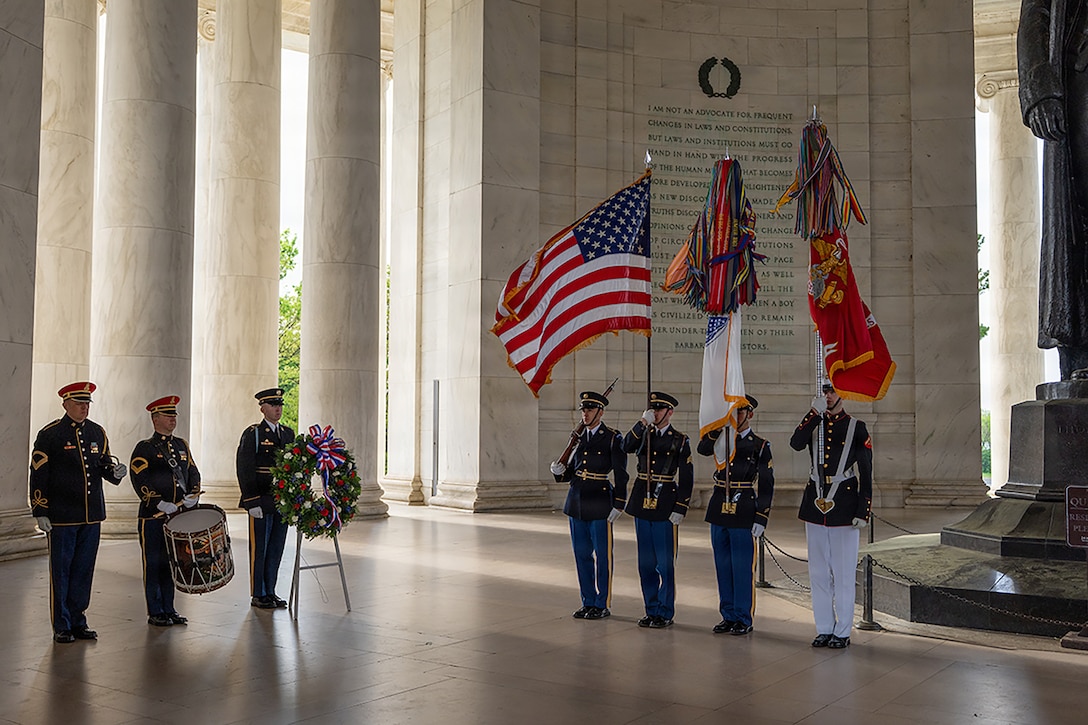 Four service members in ceremonial dress stand to the right while holding flags as fellow service members stand next to a wreath in a memorial.