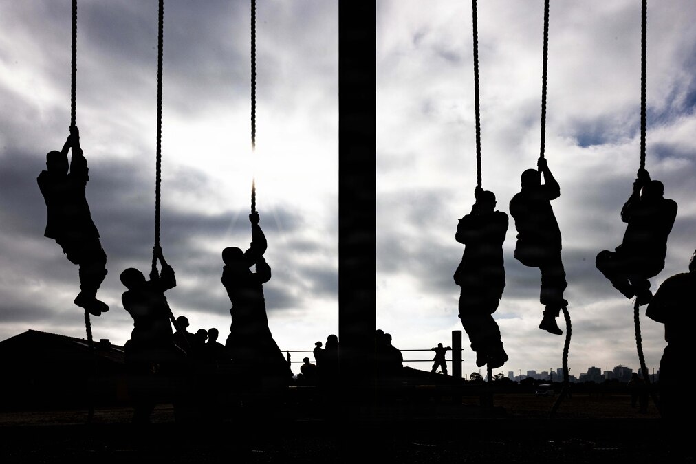 Six Marine Corps recruits climb ropes next to one another as seen in silhouette.
