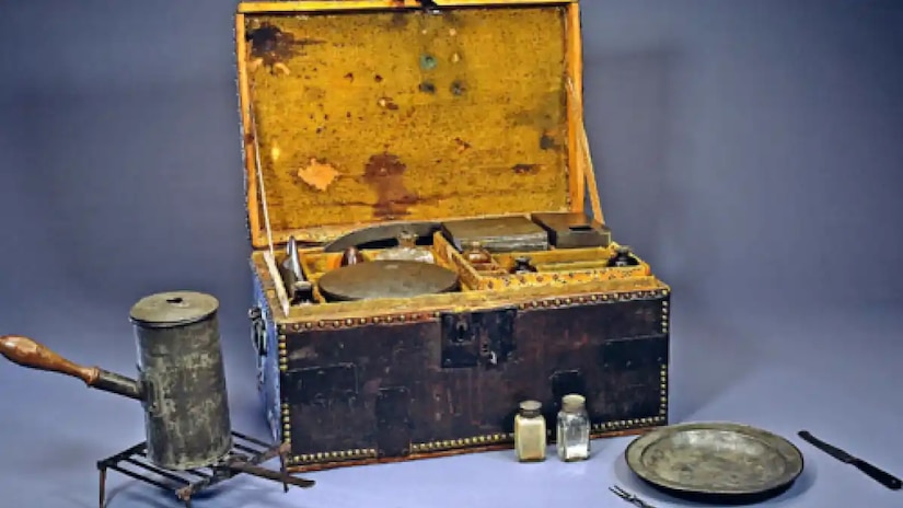A chest filled with nicknacks is displayed beside a kettle on a grill, saltshaker, plate and utensils.