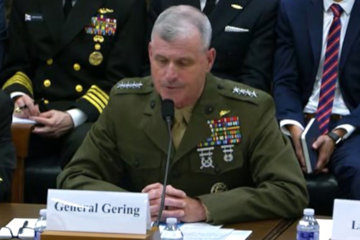 A man in a dress military uniform sits at a table and speaks into a microphone. In front of him is a card that reads, "General Gering."