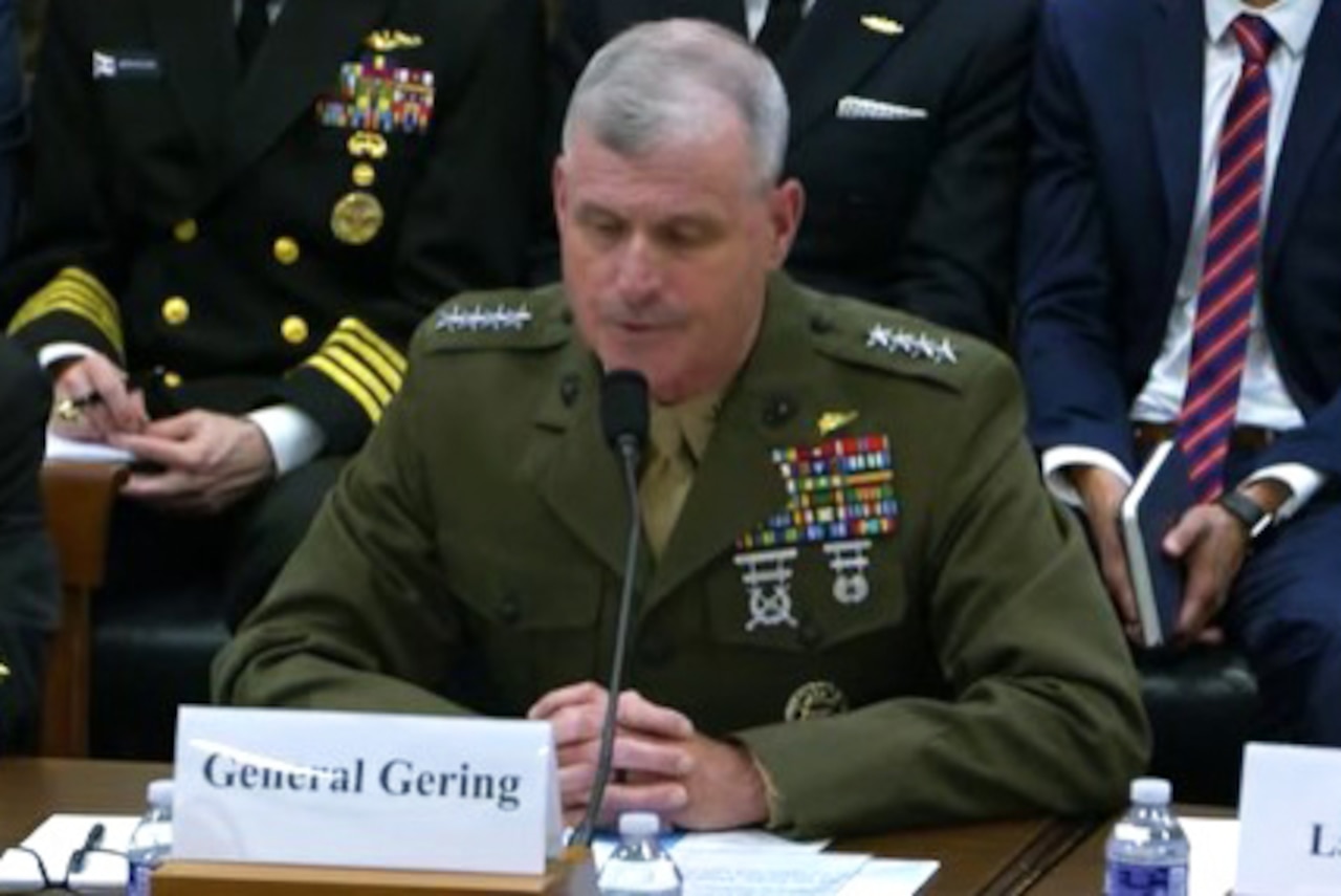 A man in a dress military uniform sits at a table and speaks into a microphone. In front of him is a card that reads, "General Gering."