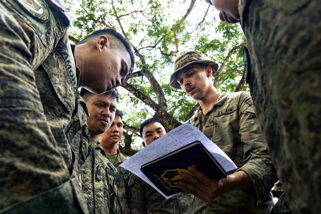 Six people in camouflage uniforms stand in a huddle outdoors and look down at a map that one of the people is holding.