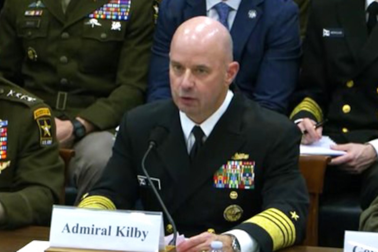A man in a dress military uniform sits at a table and speaks into a microphone. In front of him is a card that reads, "Admiral Kilby."