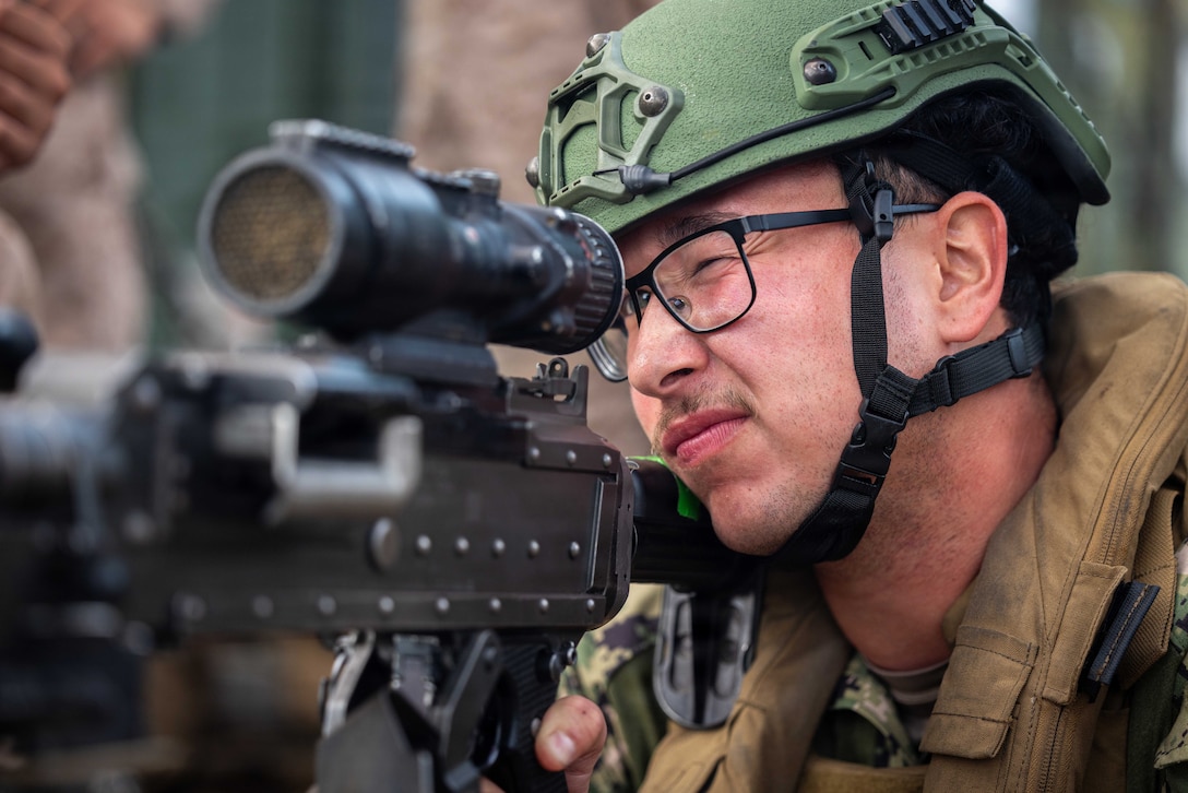A soldier holding a machine gun closes one eye and looks through a part on the gun with the other.