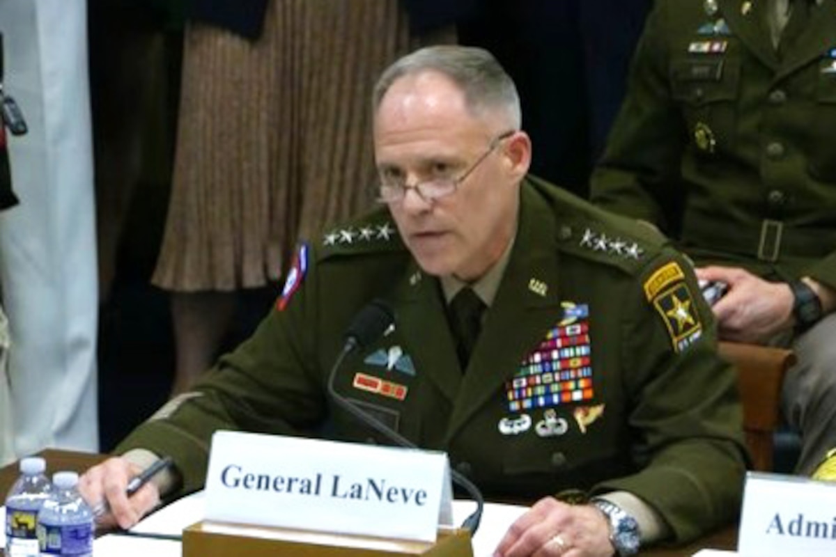 A man in a dress military uniform sits at a table and speaks into a microphone. In front of him is a card that reads "General LaNeve."