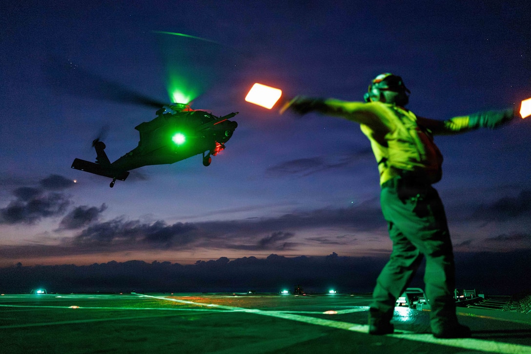 A person holding two lights stands on a ship deck at night with their arms stretched out to signal to a helicopter that is in the air.