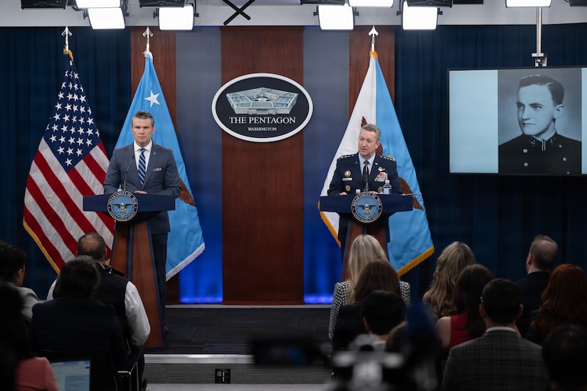 Two men, one in a business suit and the other in a military dress uniform, stand at lecterns indoors, with flags and a screen showing a person in the background.