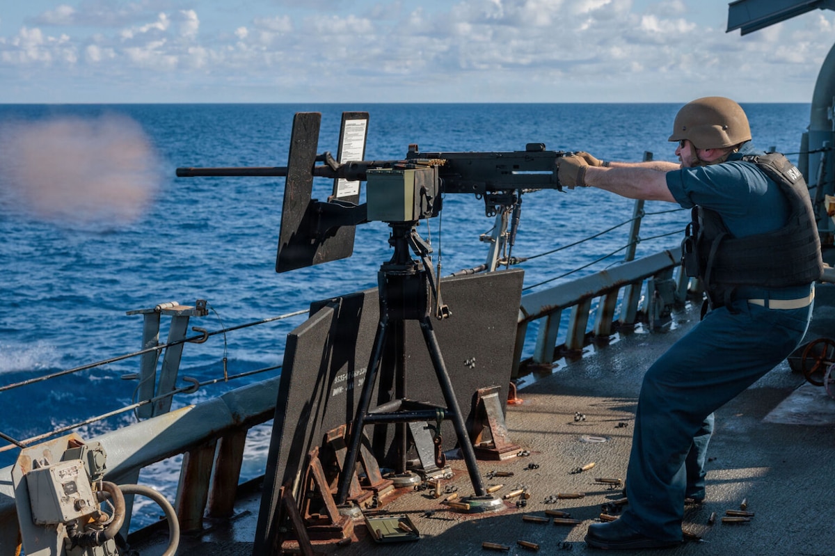 A man in a military uniform and helmet fires a machine gun into the water while sailing on a military ship.