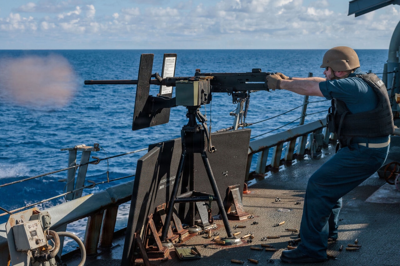 A man in a military uniform and helmet fires a machine gun into the water while sailing on a military ship.