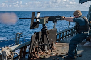A man in a military uniform and helmet fires a machine gun into the water while sailing on a military ship.