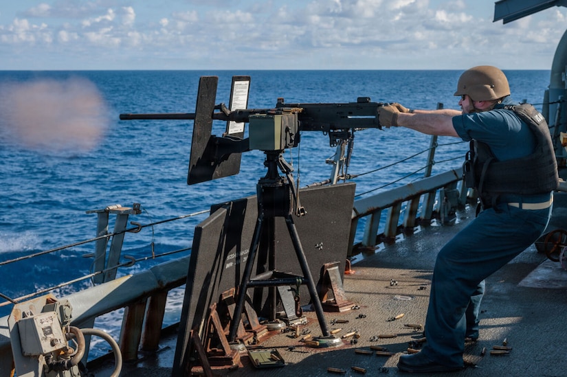 A man in a military uniform and helmet fires a machine gun into the water while sailing on a military ship.