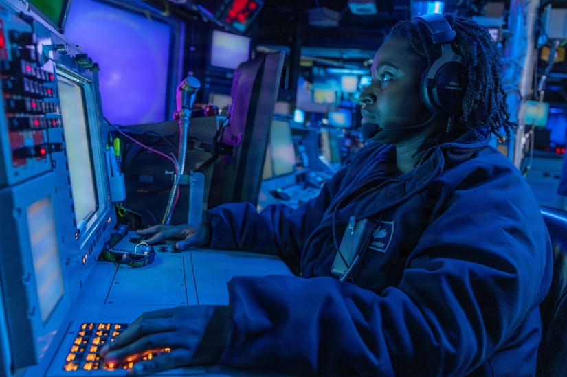A woman in a military uniform uses a keypad, while looking at a monitor in a dark room aboard a military ship.
