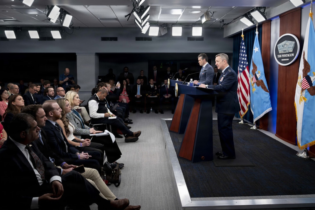 Two men stand on stage behind lecterns and speak to an audience. Behind them are an American flag and various other flags.