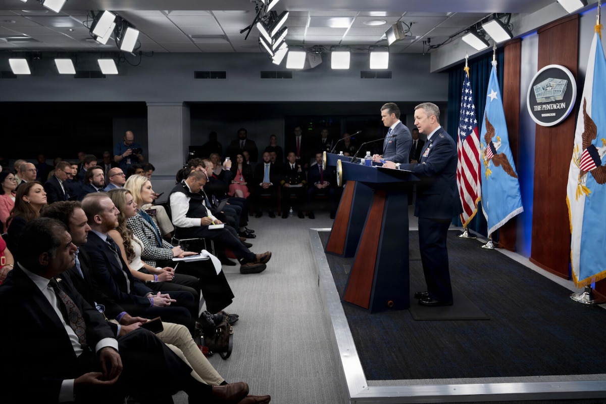 Two men stand on stage behind lecterns and speak to an audience. Behind them are an American flag and various other flags.