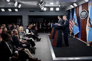 Two men stand on stage behind lecterns and speak to an audience. Behind them are an American flag and various other flags.