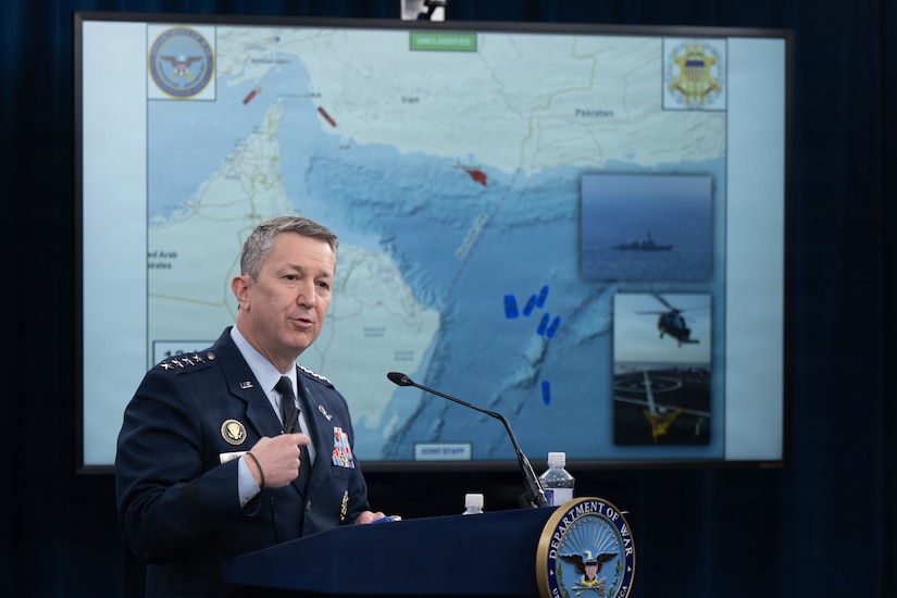 A man in a uniform stands behind a lectern. Behind him is a monitor that displays a map of the Persian Gulf and Strait of Hormuz.